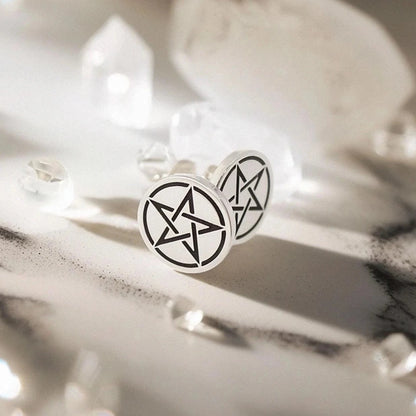A pair of silver stud earrings with pentagram symbols, displayed on a white marble surface surrounded by clear crystal points under bright, dramatic lighting.